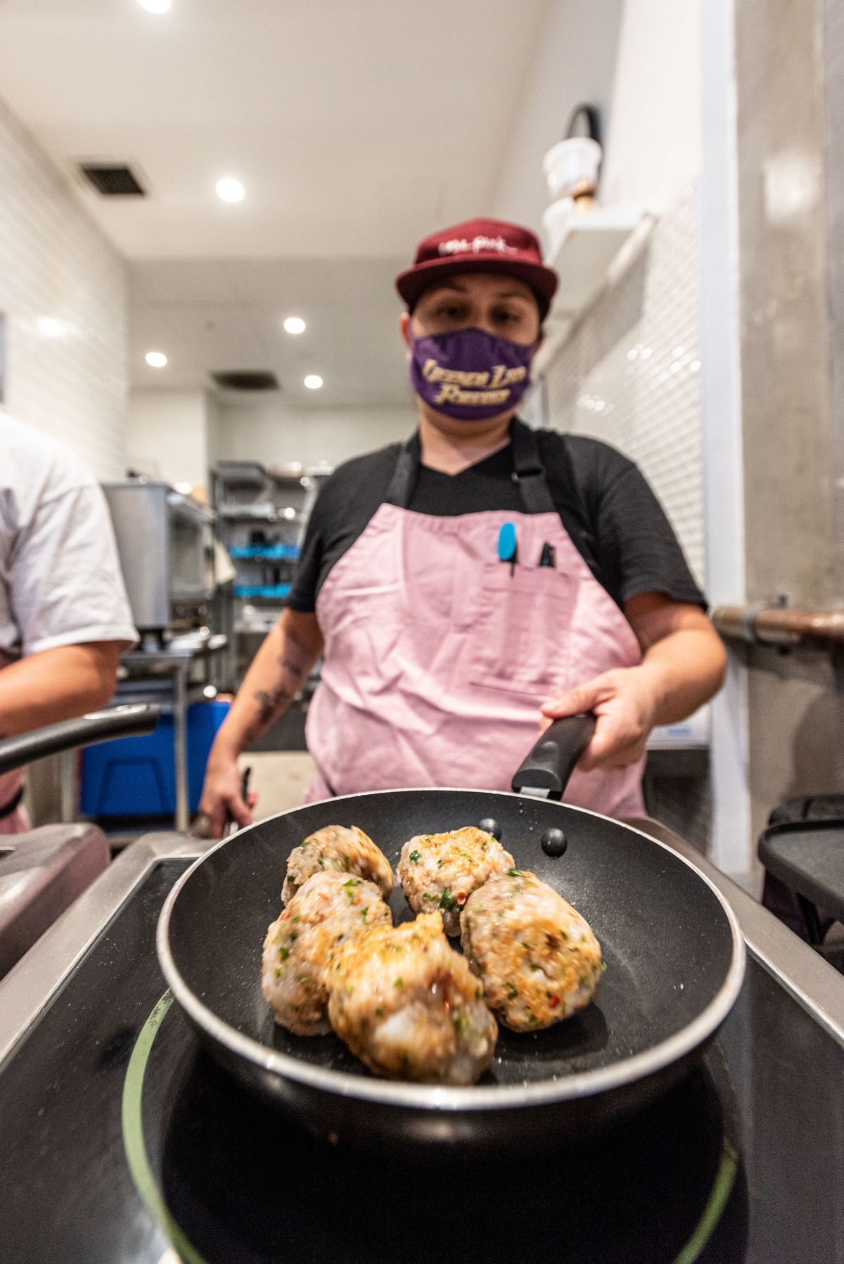 A chef in a red hat and pink apron turns meatballs in a pan.