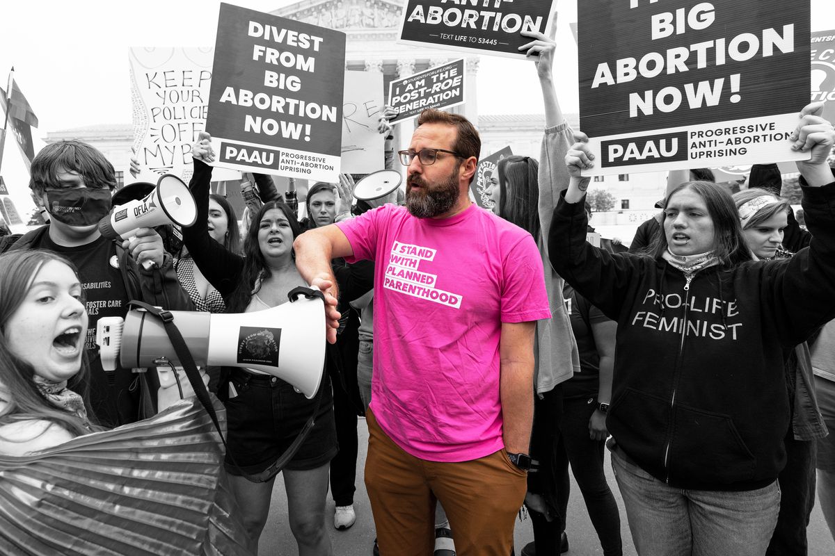 A man wearing a pink Planned Parenthood shirt puts his hand over the megaphone for an anti-abortion protestor. 