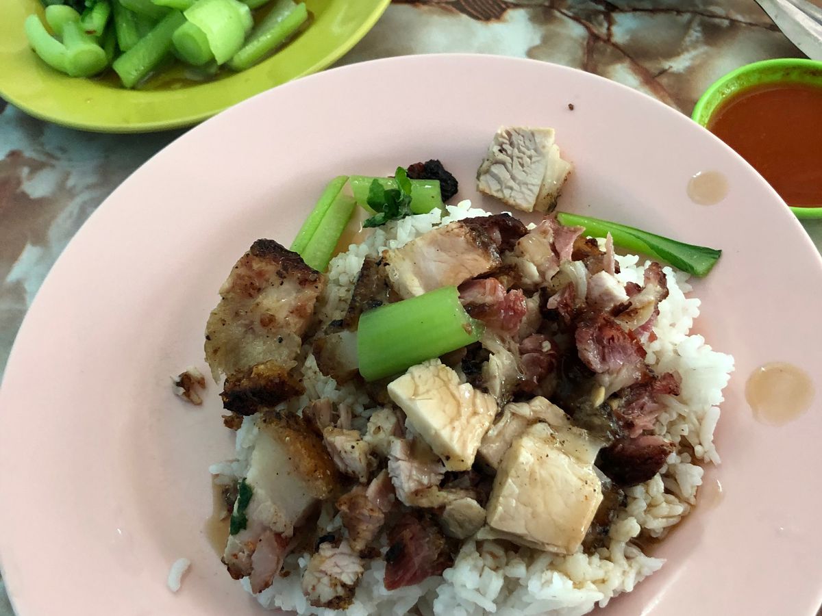 Slice of roasted pork on top of rice with greens on a pink plate beside a small plastic dish of extra greens, and two small cups of dipping sauce, on a colorful, marble-like backdrop