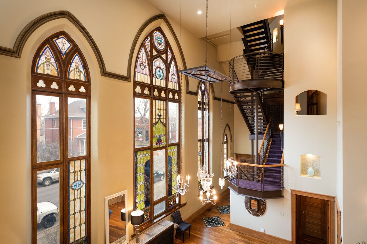 A church turned house features tall stained glass windows on one wall and very tall ceilings. 