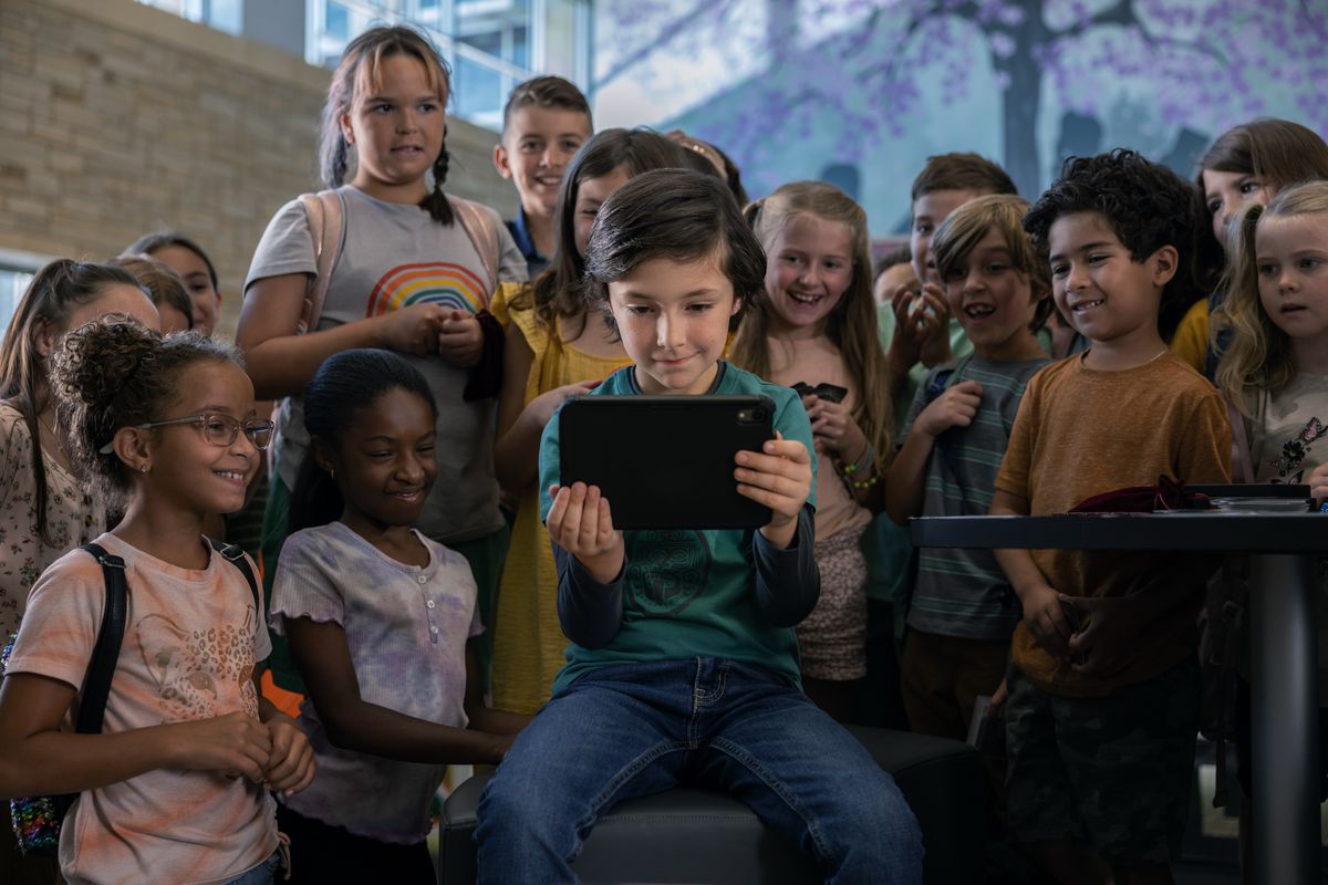 A young boy playing a game on his tablet, with a crowd of people around him to cheer him on
