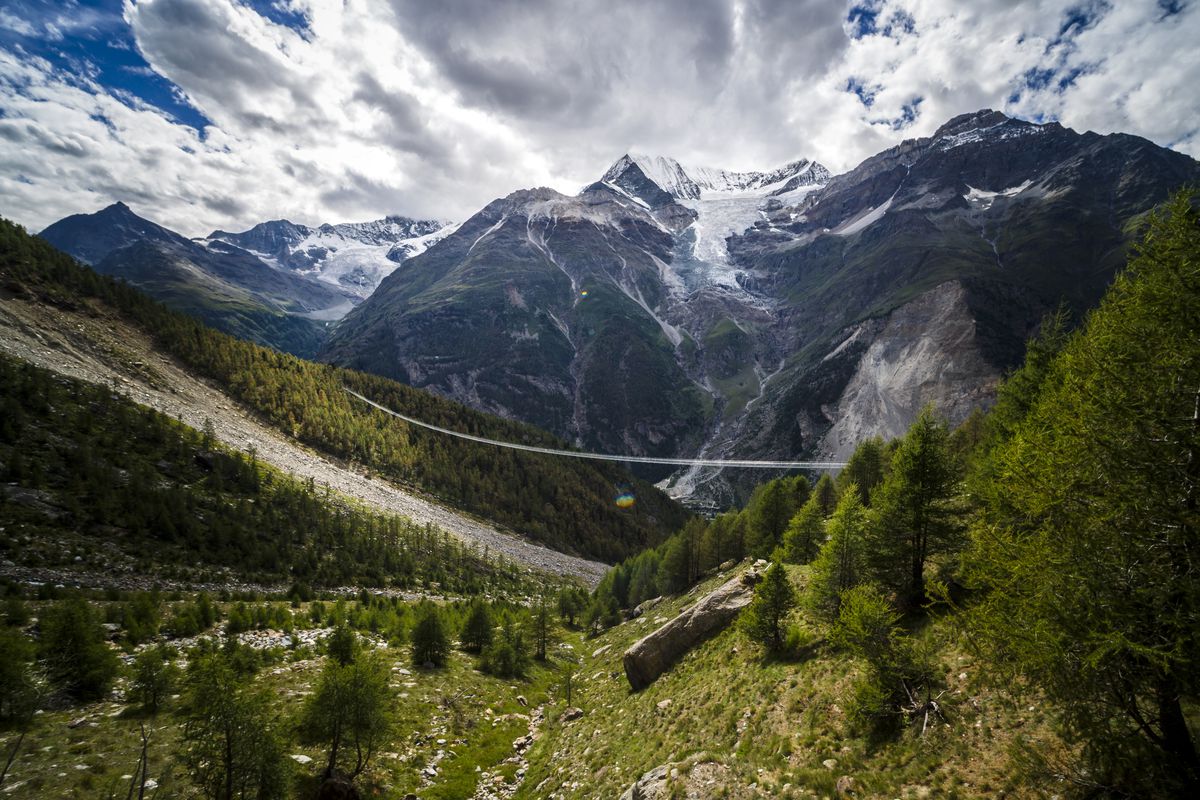 A narrow metal suspension bridge spans two sides of a valley with snowy alps in the background. 