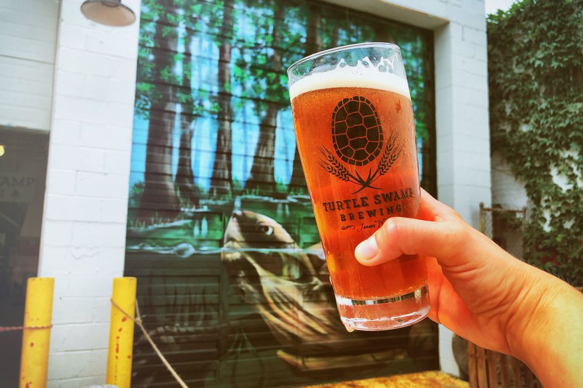 A hand holds a full glass of beer up against a mural of a turtle in a swamp