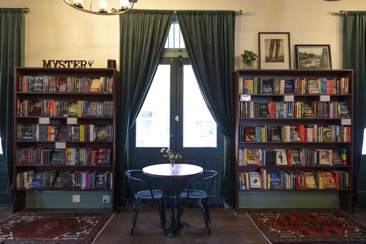 A bookstore wall with two bookshelves with books with a large window with blue curtains and a table.