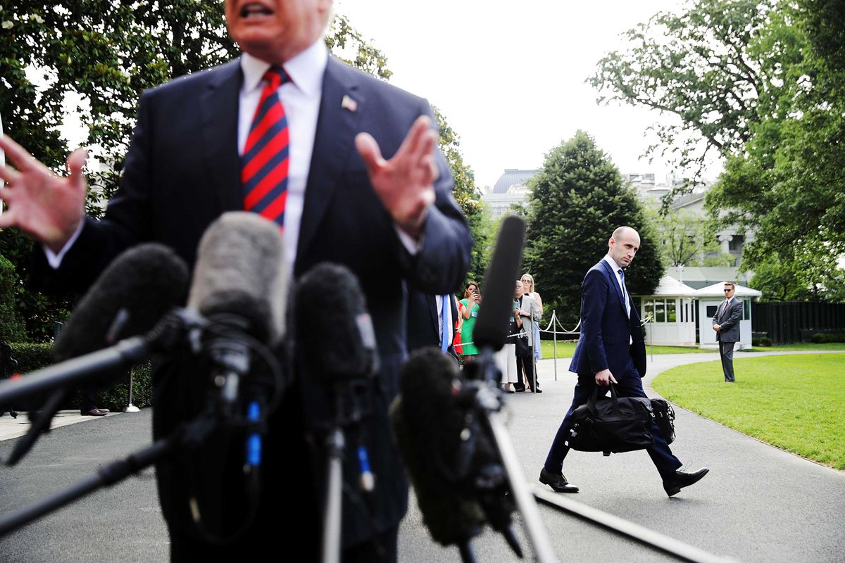 White House senior adviser Stephen Miller, in navy suit, walks behind President Trump on the White House grounds. In the foreground of the photo, Trump speaks to reporters, gesturing above a cluster of microphones.