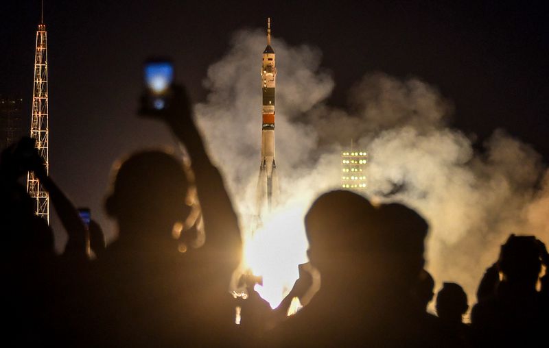 A Soyuz rocket launching, with its engines firing, as people on the ground take photos and watch.