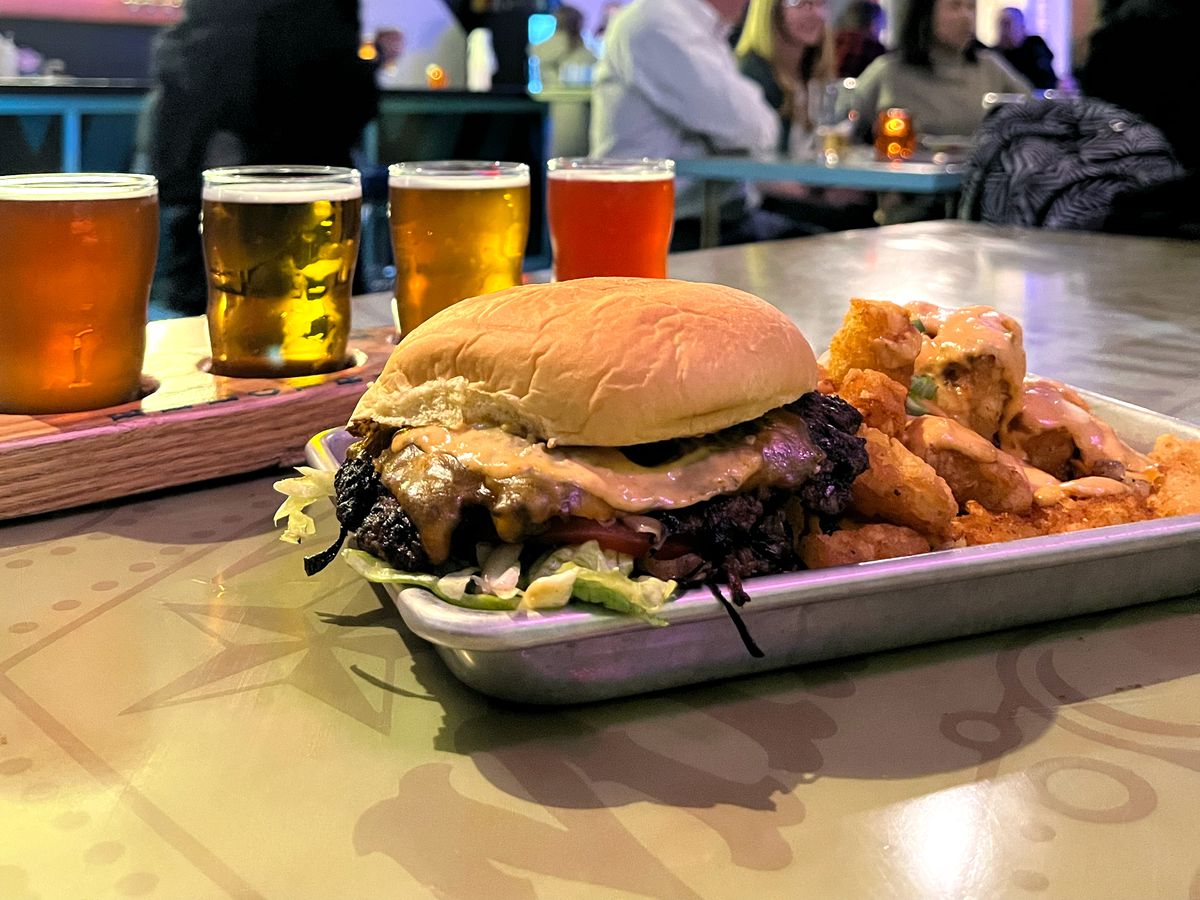 A wide flat burger topped with sauce and lettuce, on a quarter sheet with fries, served beside a flight of beers.