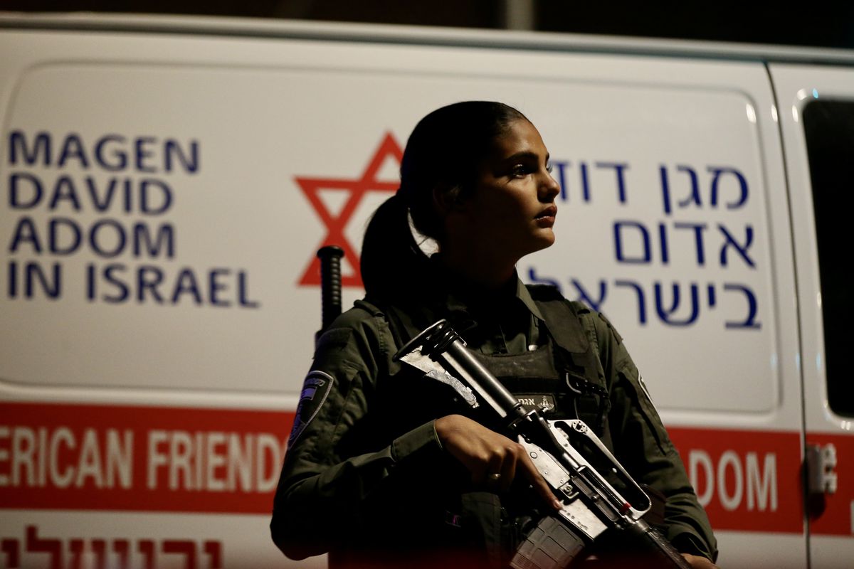 A woman Israeli police officer wearing a camouflage uniform holds a military-style rifle and looks to her left. Behind her is an Israeli ambulance.