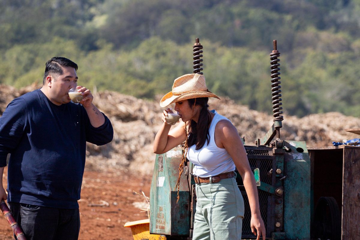 Chris Kajioka (left), in a dark blue shirt and black jeans, and Padma Lakshmi (right), in a white tank top, green cargo pants, boots and a cowboy hat, drink sugar cane juice on a sugar cane farm.