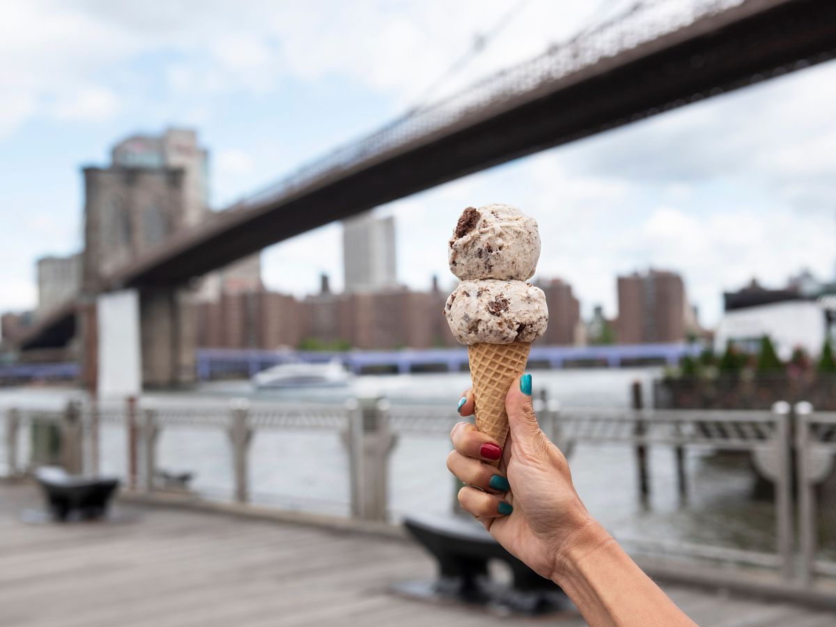 A hand holds an ice cream cone with two scoops near the waterfront in NYC. 
