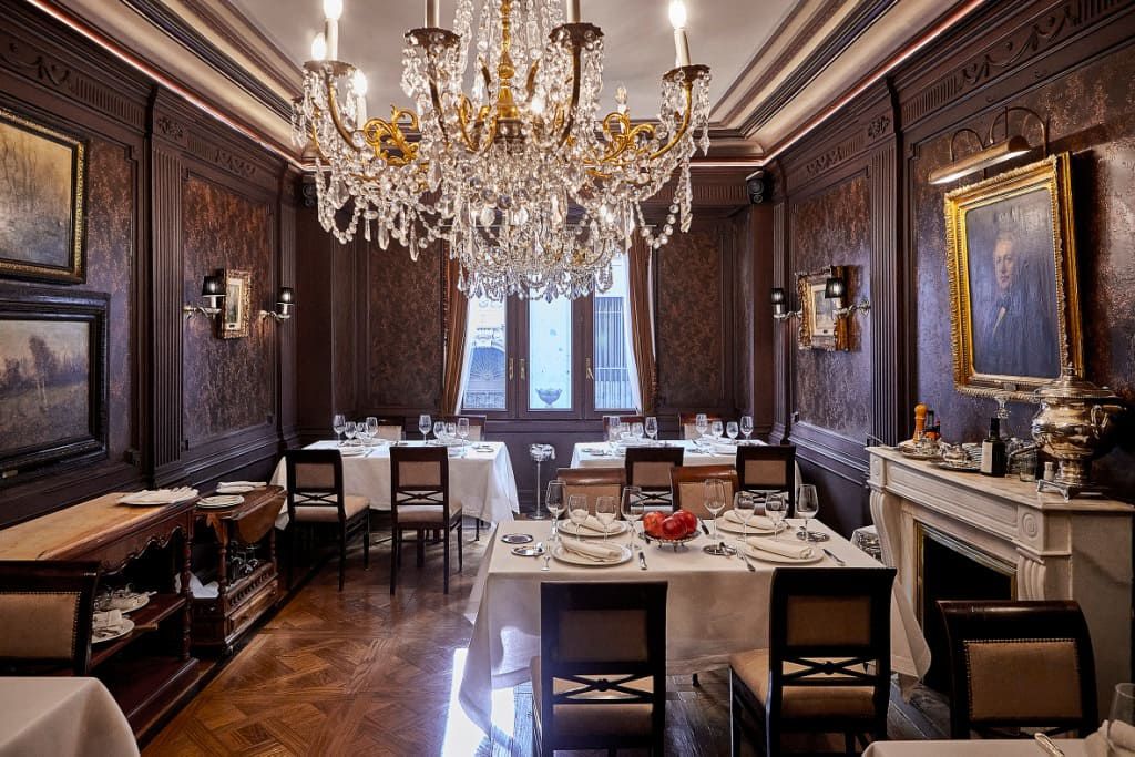 A formal dining room with dark wood walls, crystal chandelier, white tablecloths, and classical art. 