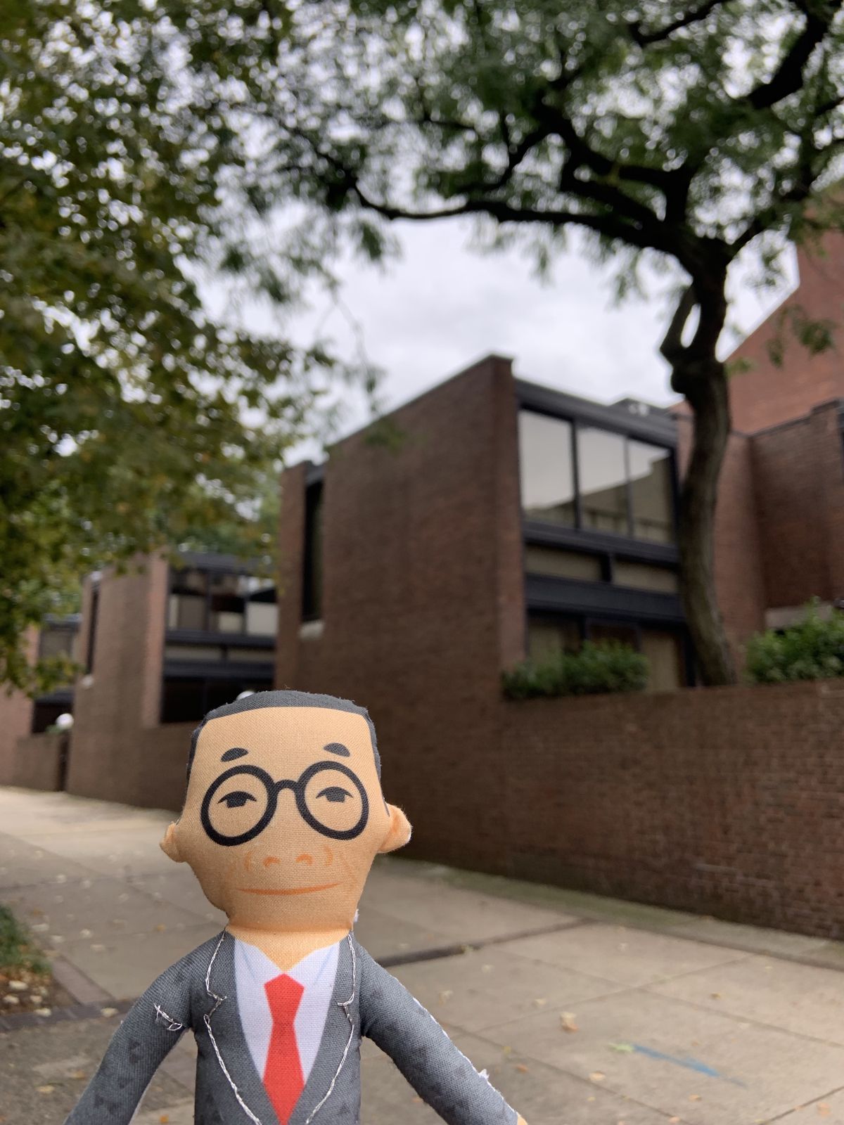 A small doll showing a bespectacled man in front of a brick and glass building.