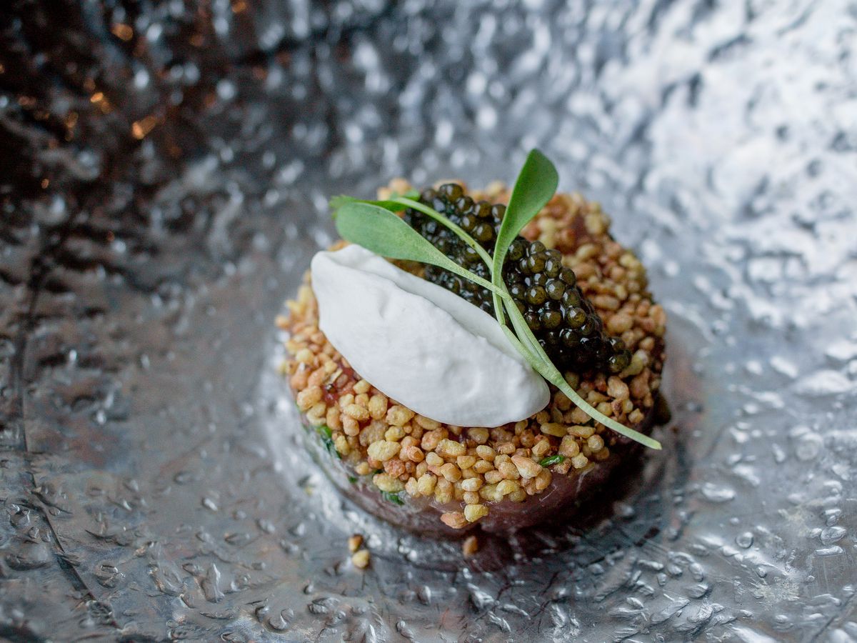 A shiny black textured dish holding a small circular patty of tuna topped with buckwheat, a collop of cream, a pile of caviar and an herb sprig for garnish