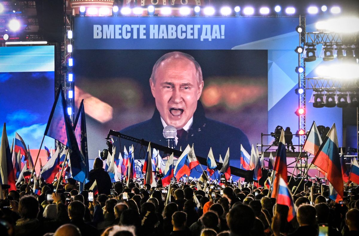 Russian President Putin appears on a huge screen in front of a crowd carrying tri-color flags.