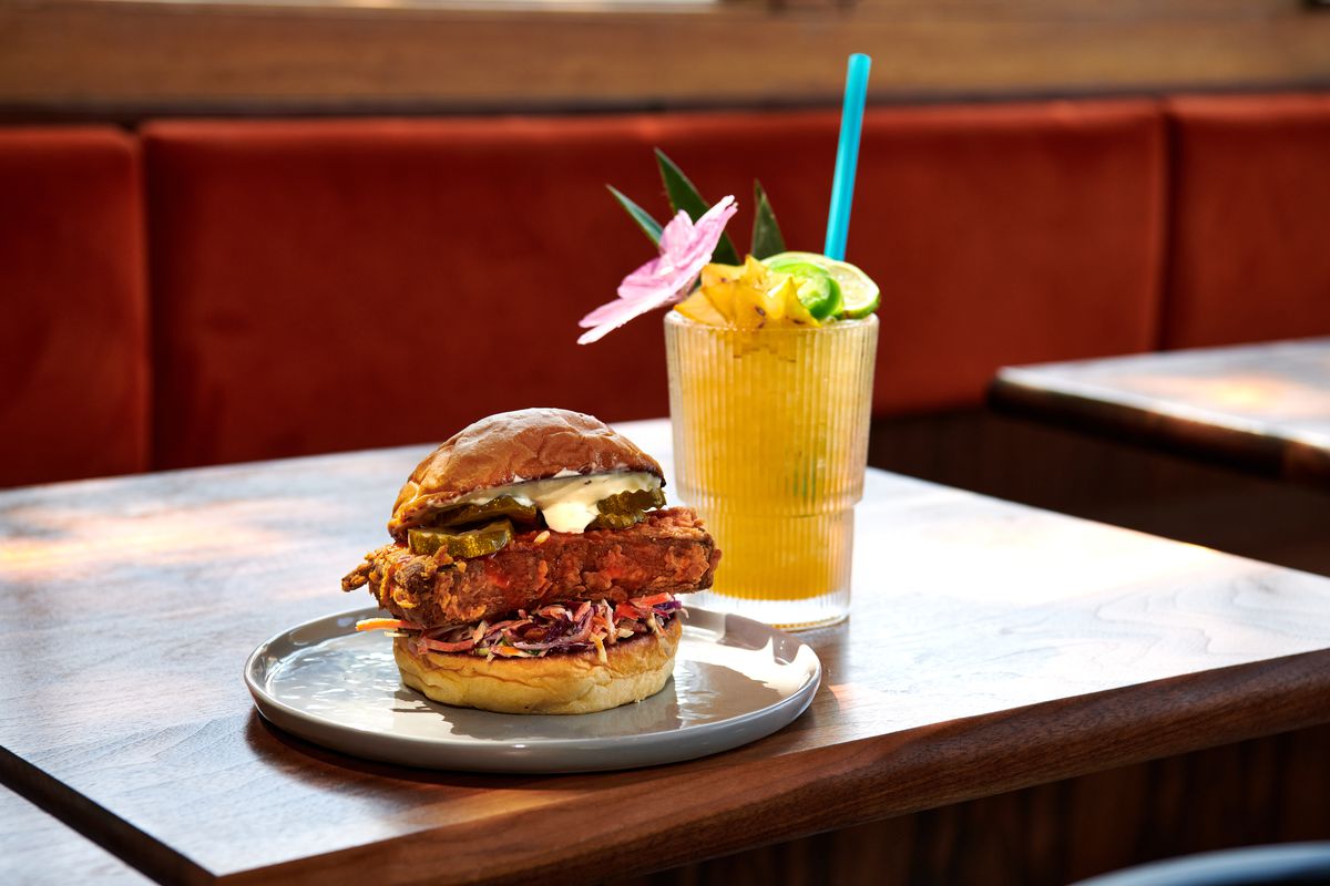 An orange cocktail with tropical fruit garnishes and a straw sits next to a fried chicken sandwich at Jojo.