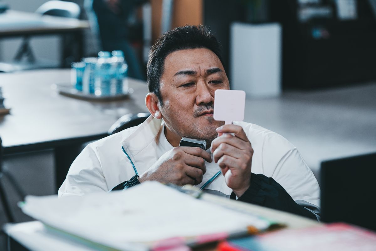 Ma Dong-seok holds up a handheld mirror while shaving his face at his desk in The Roundup: No Way Out.