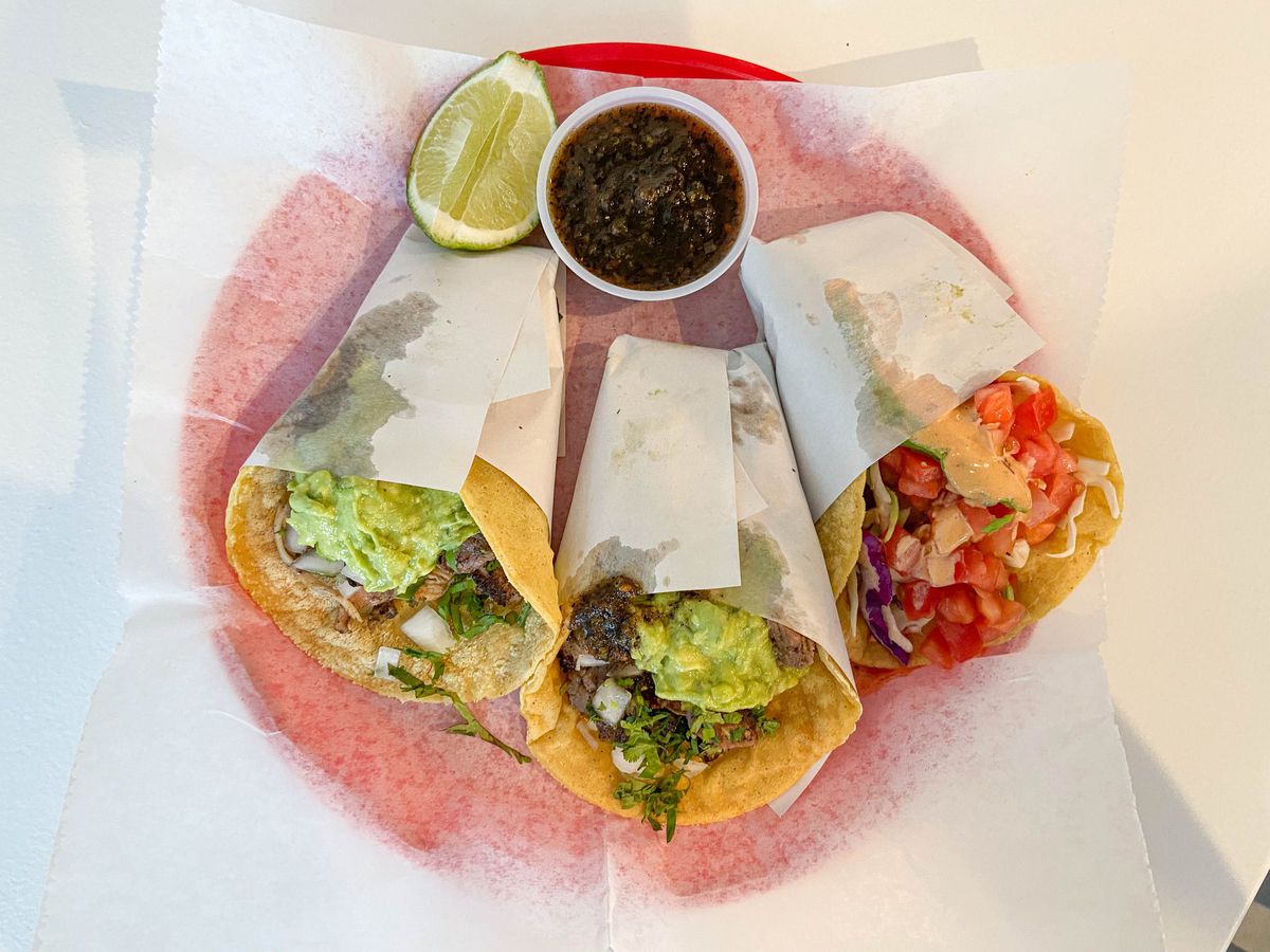 Three different tacos, all wrapped in paper and served side by side on a plate with salsa on the side.