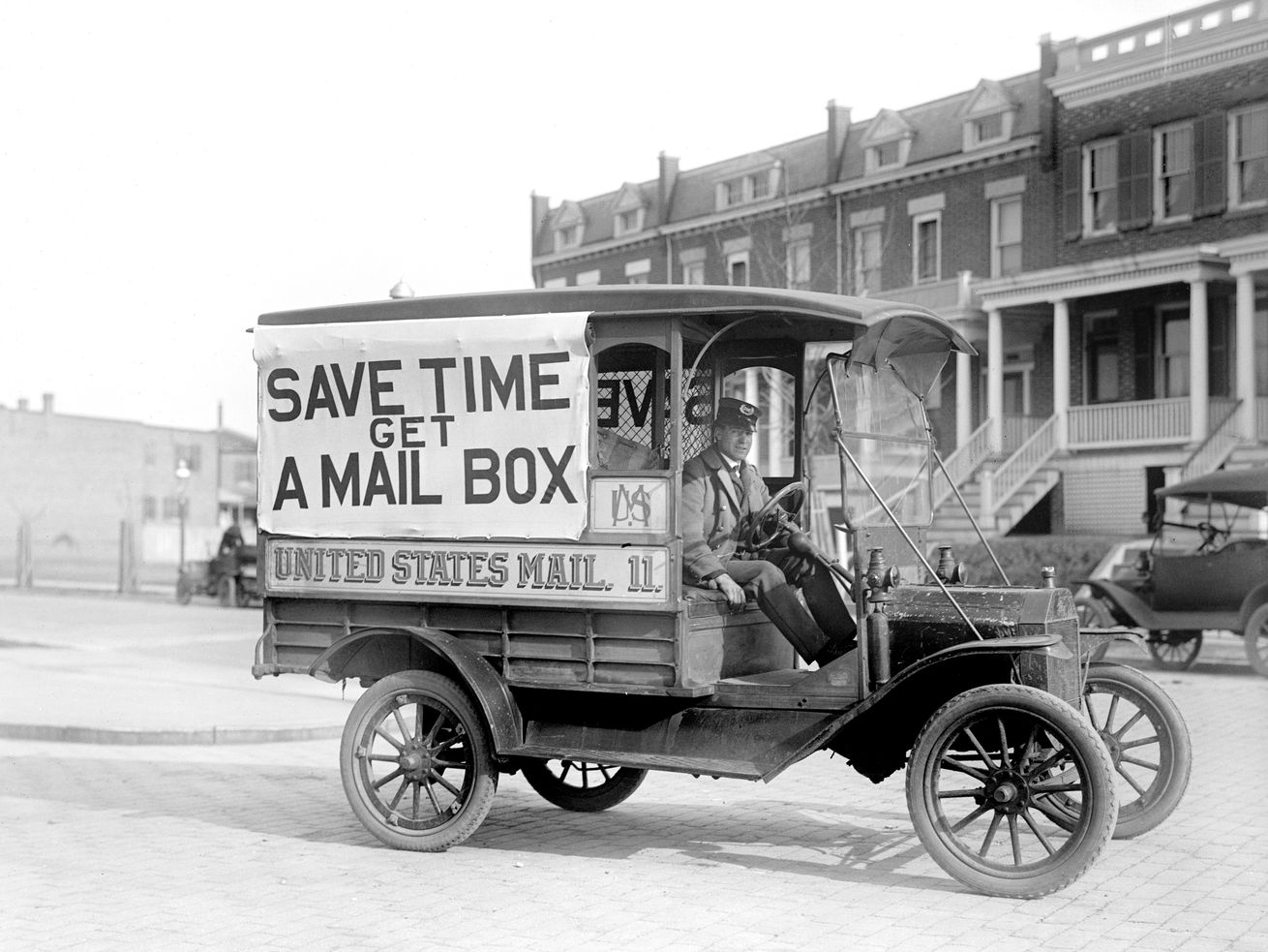 A car resembling a Model T Ford carries a sign on its side that reads, “Save time, get a mail box. United States mail.