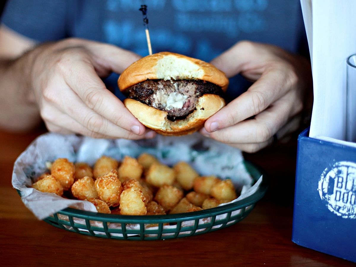A person holding a burger with a bite out of it, cheese is oozing out of the center, there is also a basket of tater tots on the table. 