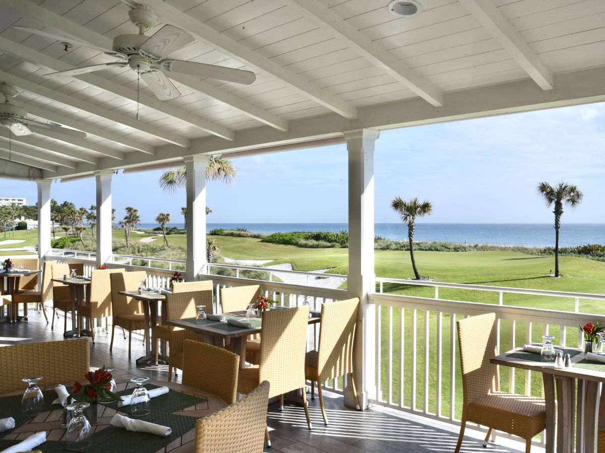 This is a picture of the outside dining porch at Al Fresco in Palm Beach. There are tables and chairs on the porch, and a painted white wood ceiling. In the background is a golf course and beyond is the Atlantic ocean.