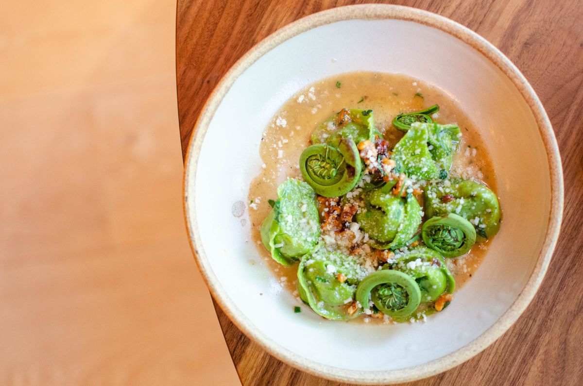 Overhead view of a bowl of green filled pasta tossed with fiddleheads, served in a thick pool of a brown butter sauce.
