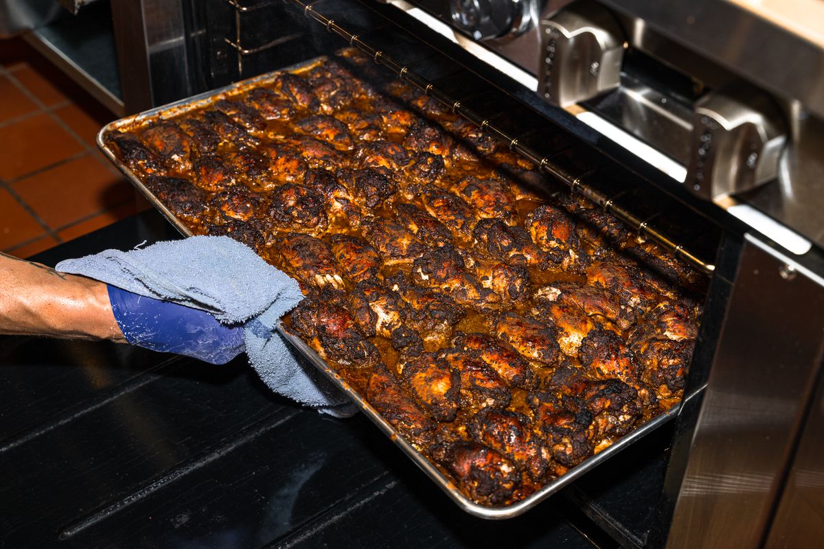 A person pulls a large tray filled with jerk chicken out of an oven.