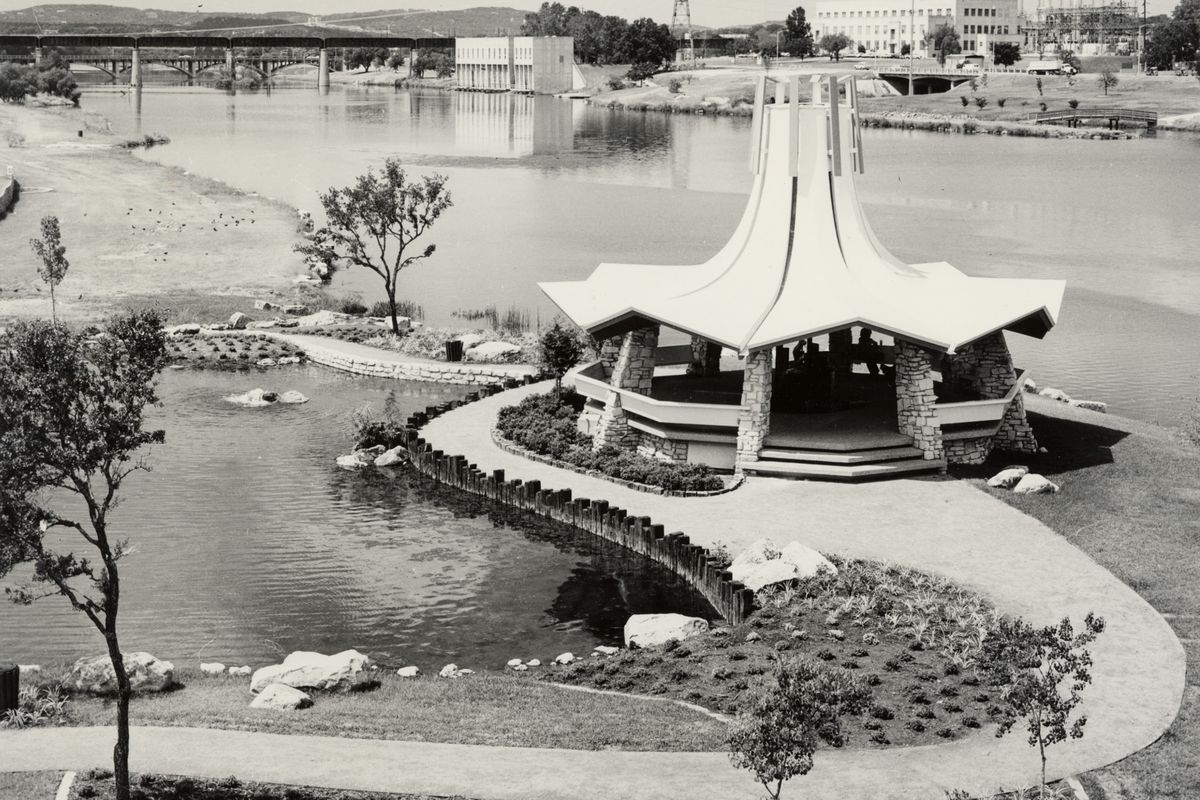 A black and white photo of a gazebo on a jetty-type land mass with a river behind it. The building has a zigzagged roof and slopes dramatically upward in a conical shape. The other shore of the lake has a few buildings on it and is in the background, along with two bridges in the distance.