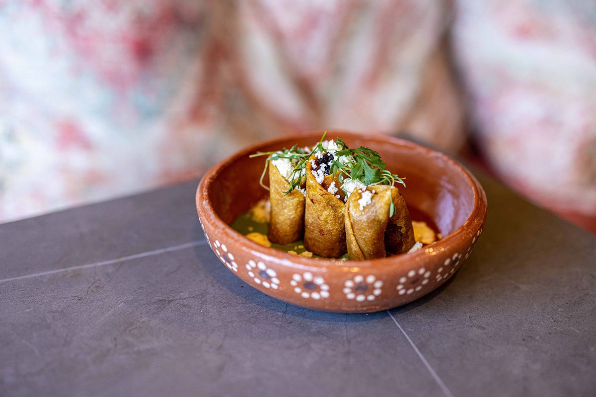 A trio of stand-up taquitos served in a clay pot with designs, on a slate table at a restaurant at daytime.