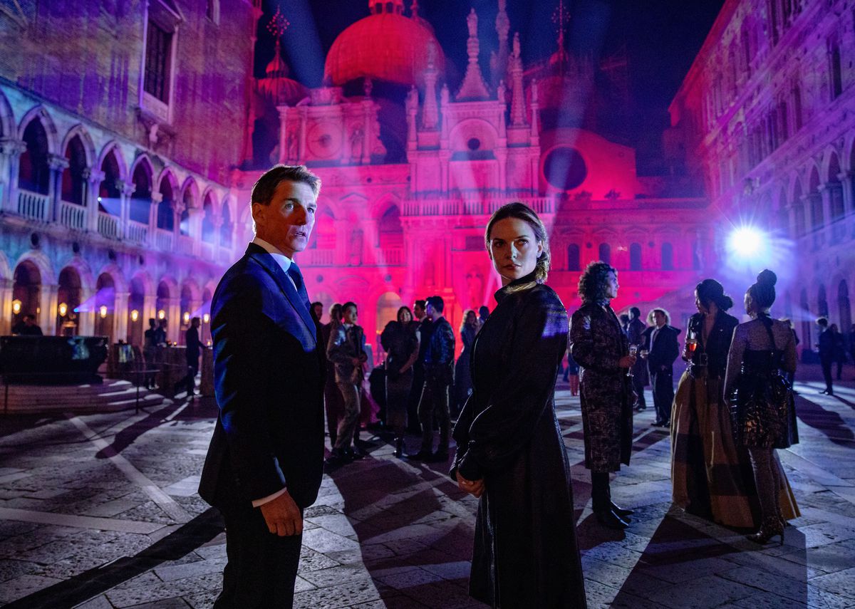Tom Cruise and Rebecca Ferguson wear formal clothing and stand near a crowd in front of a pink and purple-tinted building in Mission: Impossible Dead Reckoning — Part One.