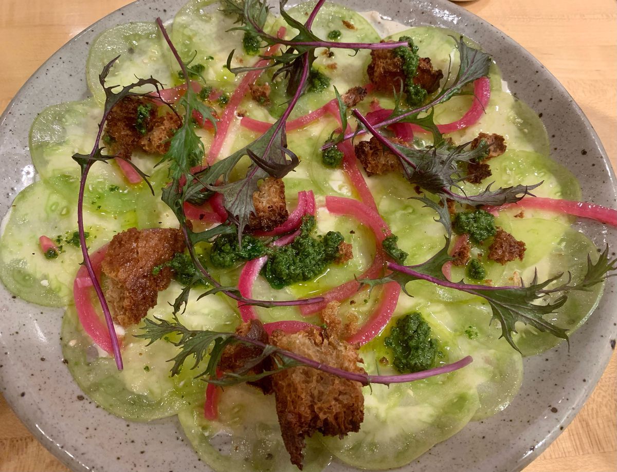 A round plate covered in thinly-sliced green tomatoes topped with croutons and pickled onion.