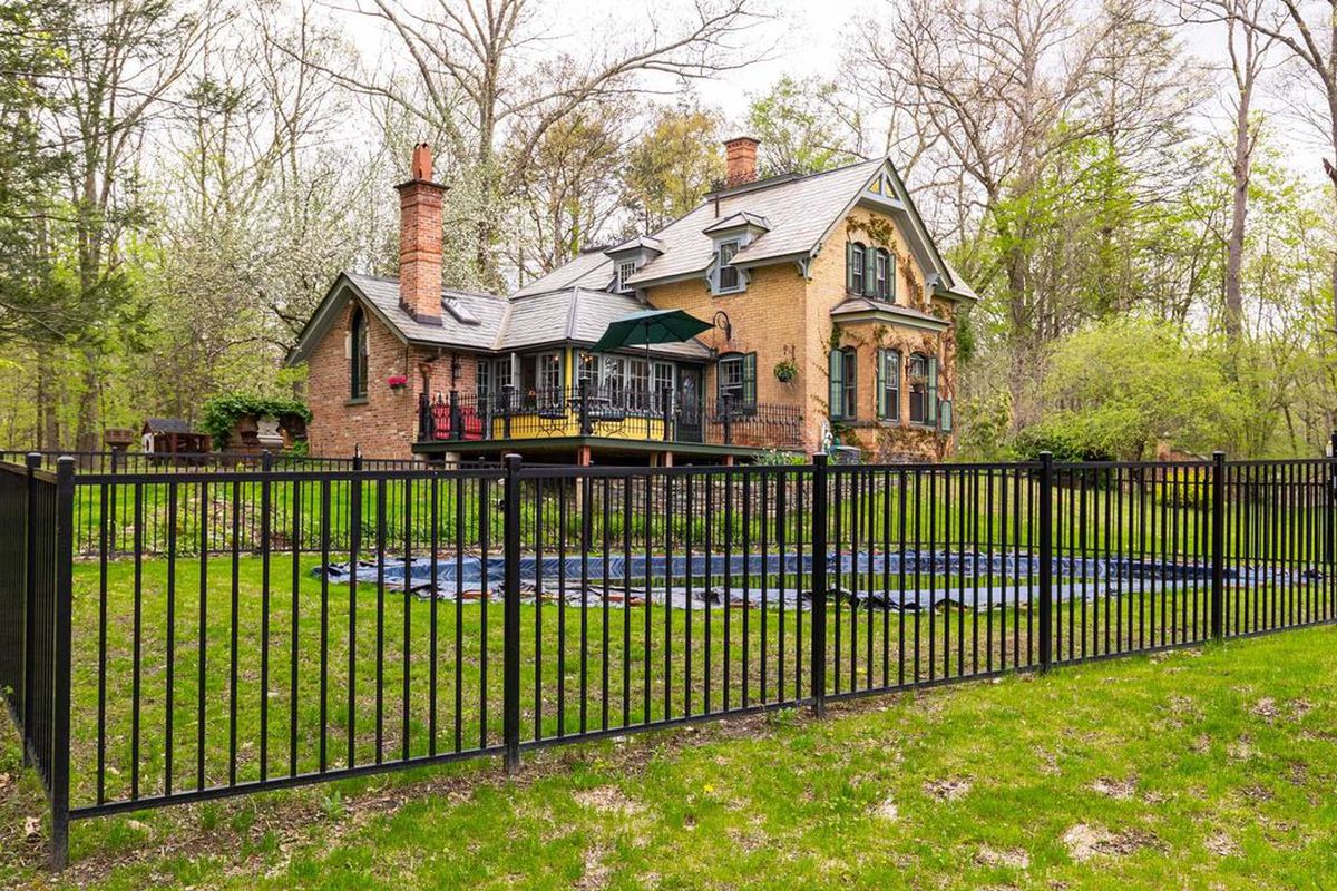 A two-story brick building sits behind an iron gate in a forested and green grass area. 