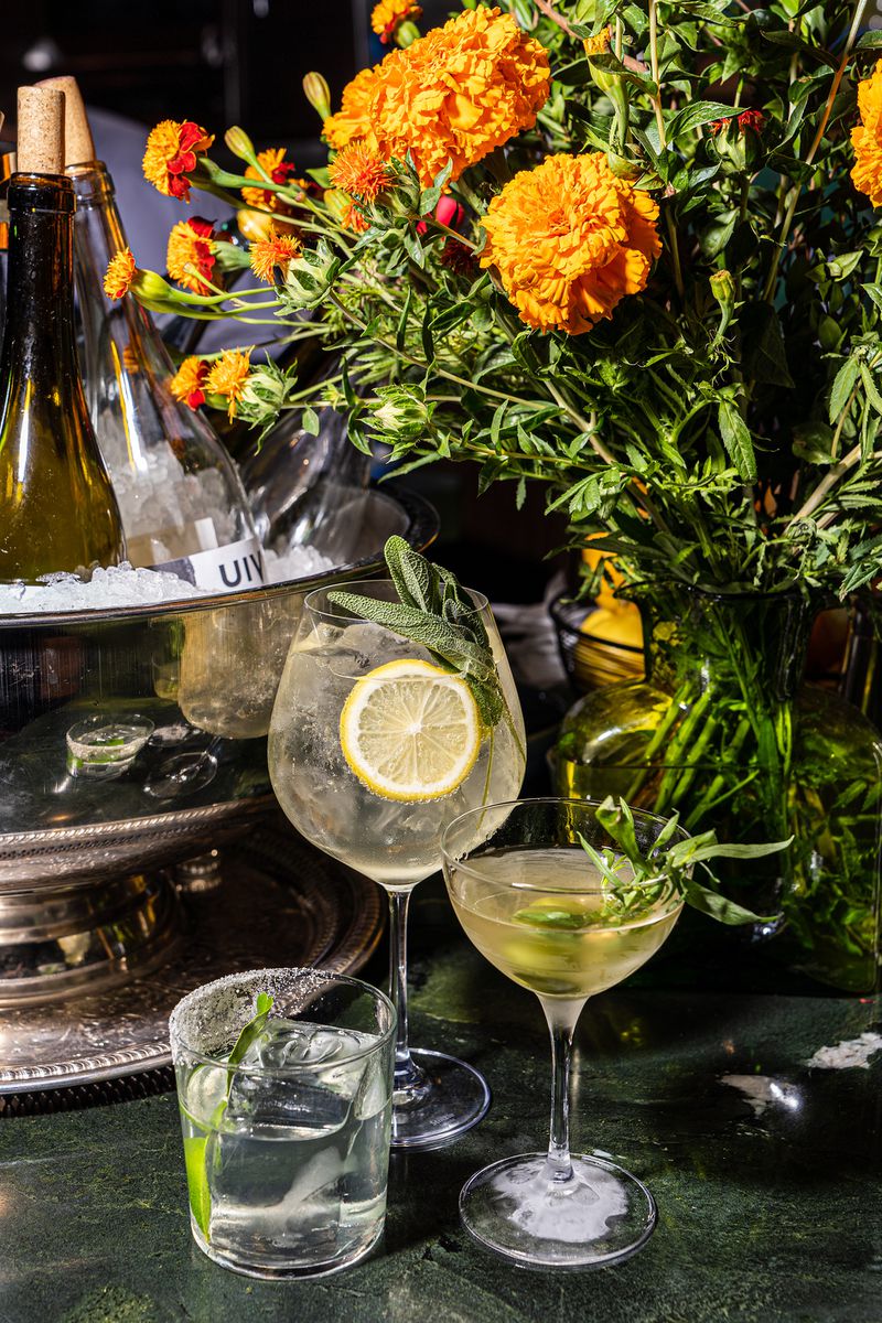 A trio of cocktails, including a gin and tonic garnished with lemon and sage, sits near a bouquet of orange flowers. 