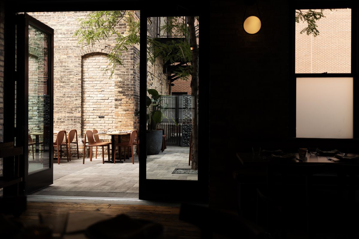 A photo from inside a darkened restaurant interior through wide double doors that open onto a sunny patio in a brick alleyway. 