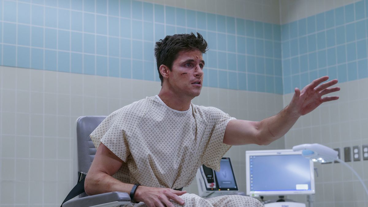 Robbie Amell, wearing a loose hospital robe and sporting a cut across one cheek, sits in a chair surrounded by medical equipment and reaches for something offscreen.