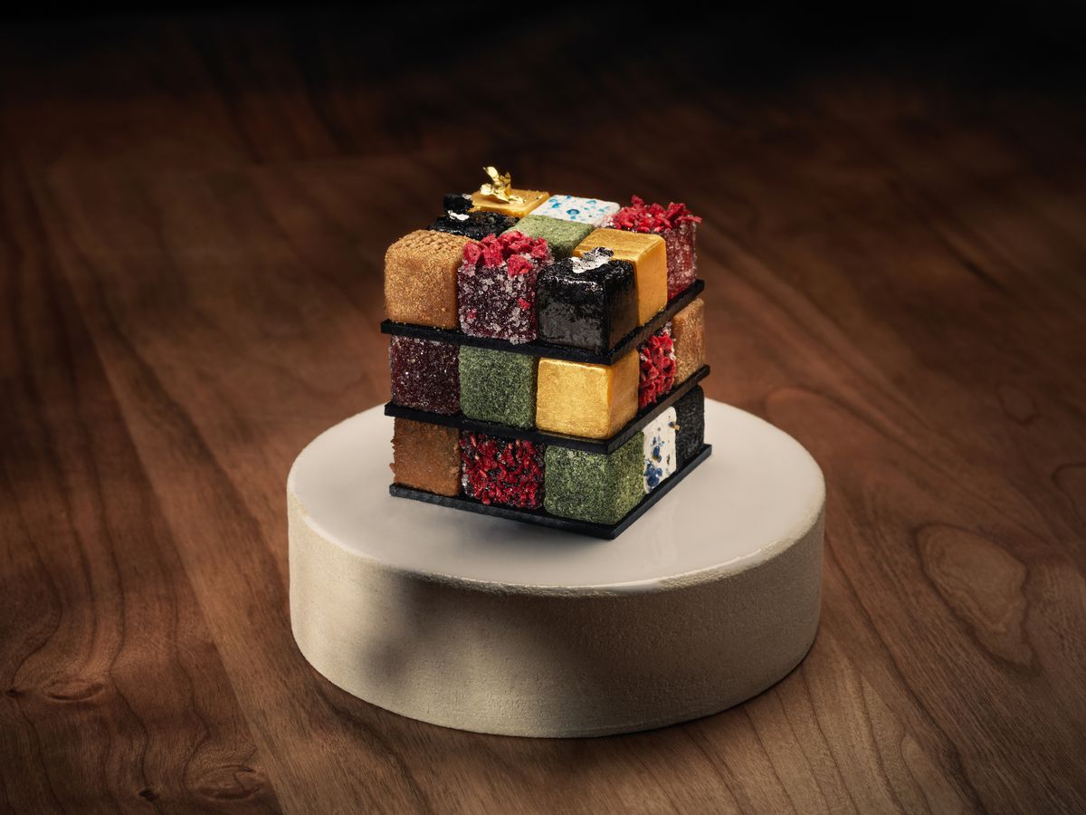 A rubik’s cube plated dessert on a white pedestal, on a wooden table.