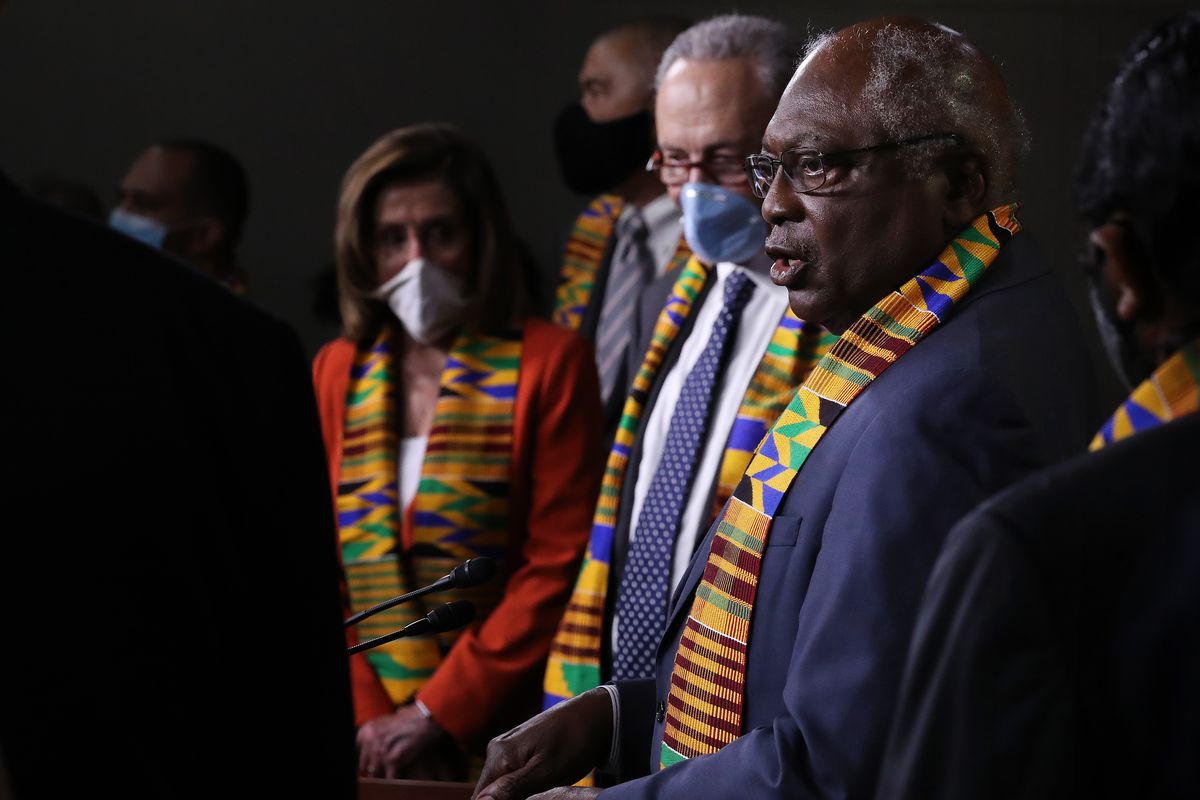 Clyburn, in a blue suit, and with a kente cloth stole around his neck, speaks as Schumer — in a navy suit — and Pelosi — in a red suit — look on. Both Pelosi and Schumer are wearing masks, and both also have on kente cloth stoles.