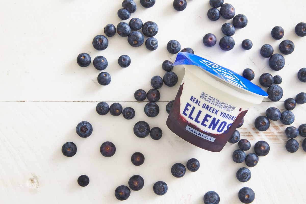 A container of Ellenos Greek Yogurt in blueberry, surrounded by loose blueberries.