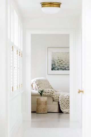 Una habitación al final de un largo pasillo blanco con un sofá blanco, una manta blanca gruesa y una mesa auxiliar de tocones de madera natural. 