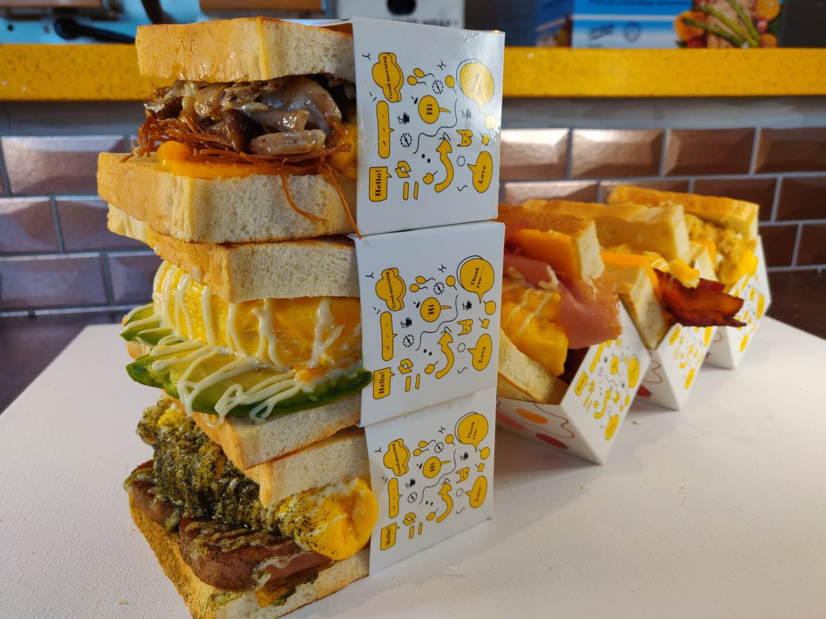 Three sandwiches on fluffy milk bread, oozing with cheese, overflowing with meat, stacked in cardboard sleeves beside paper boats of french fries