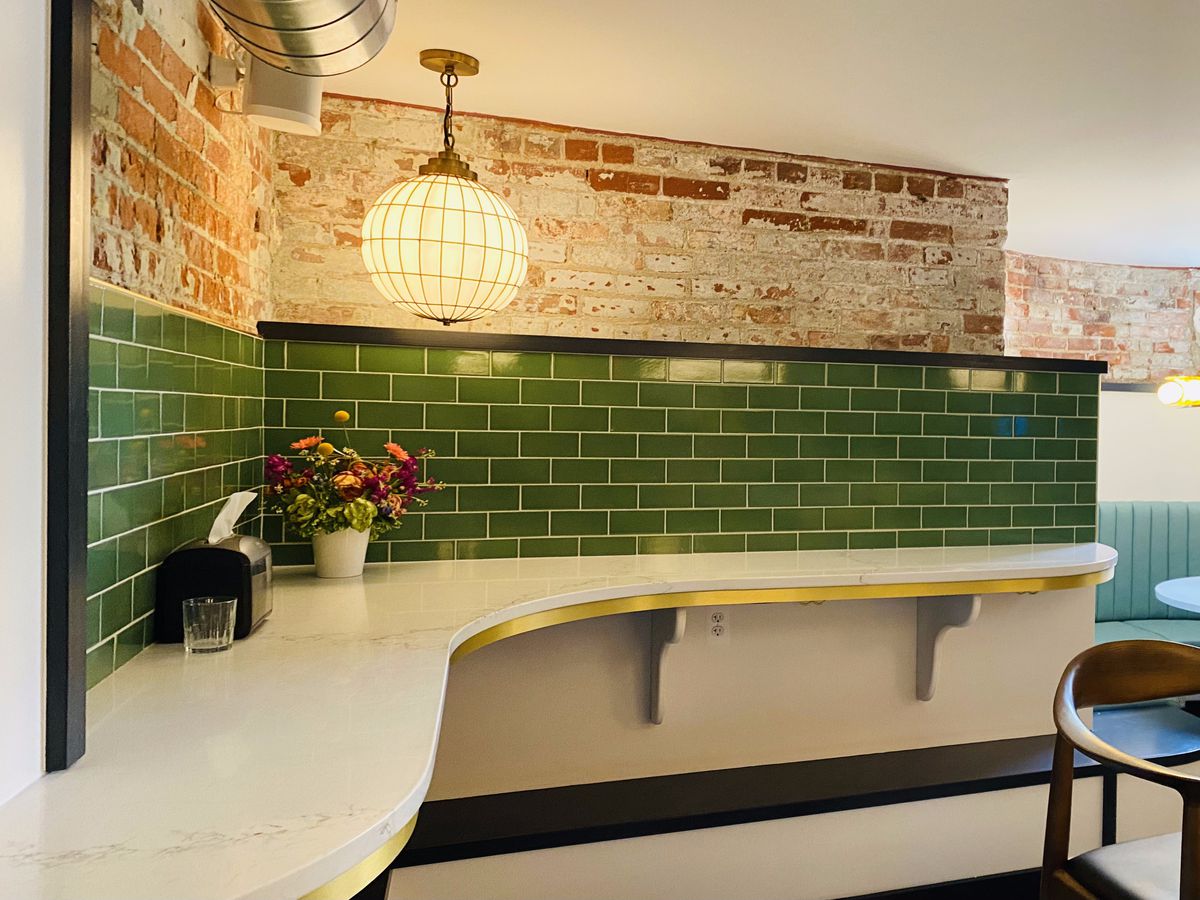 A standing bar with green tiles in the background and exposed brick wall with a flower, glass, and napkin dispenser on top.