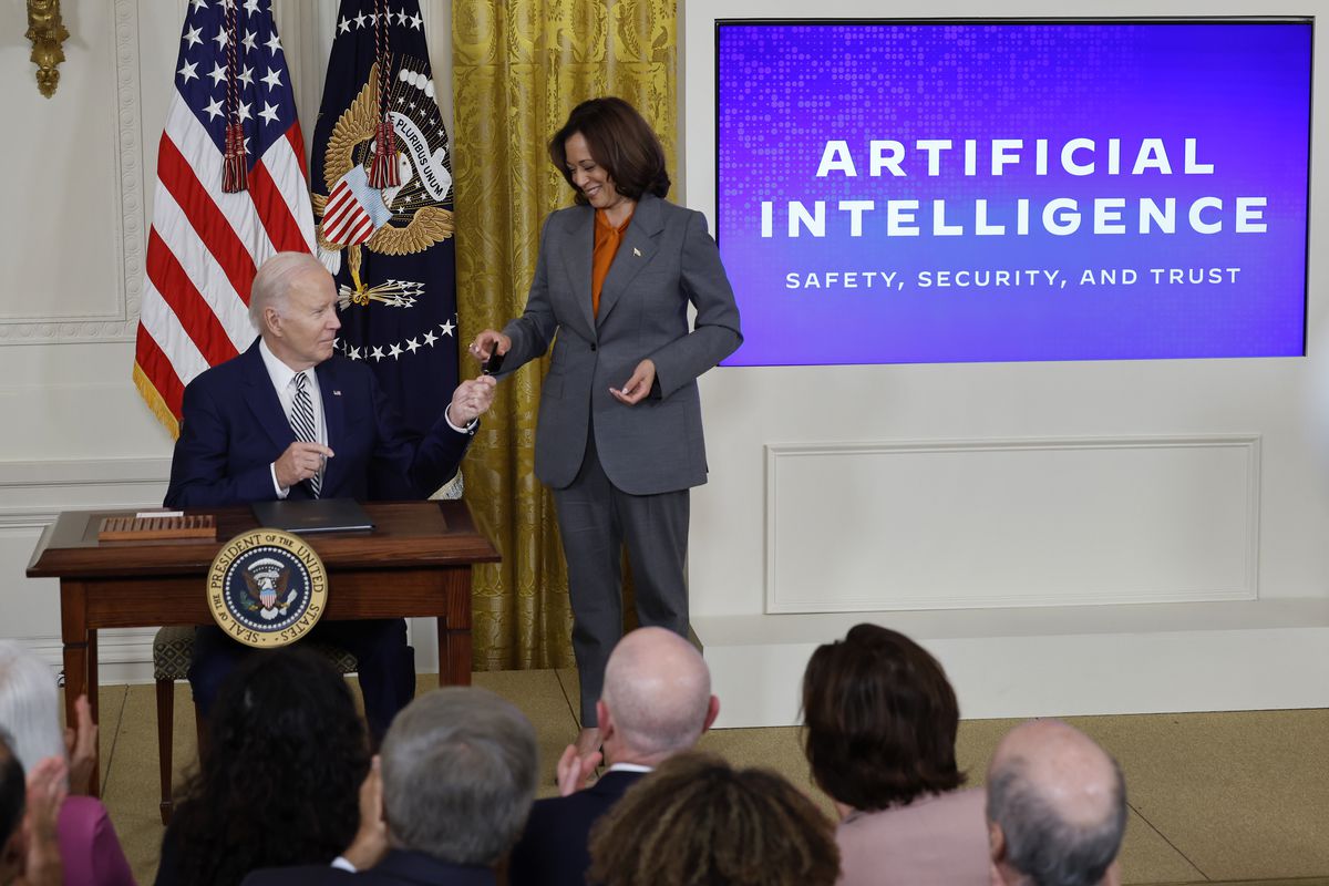President Joe Biden, sitting at a desk, hands his pen to Vice President Kamala Harris, standing, while a presentation on AI is projected on a screen next to them.