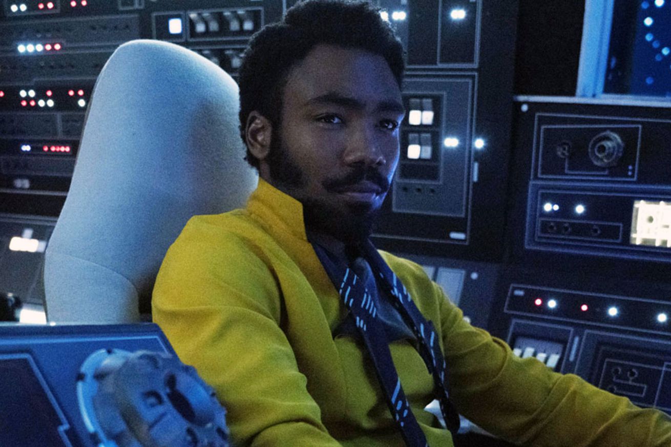 A stylish man in a yellow shirt sitting in the cockpit of a spaceship surrounded by an array of controls.