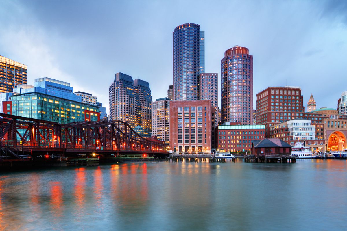 A view of the Boston skyline, featuring Financial District skyscrapers, water, and a cloudy gray sky
