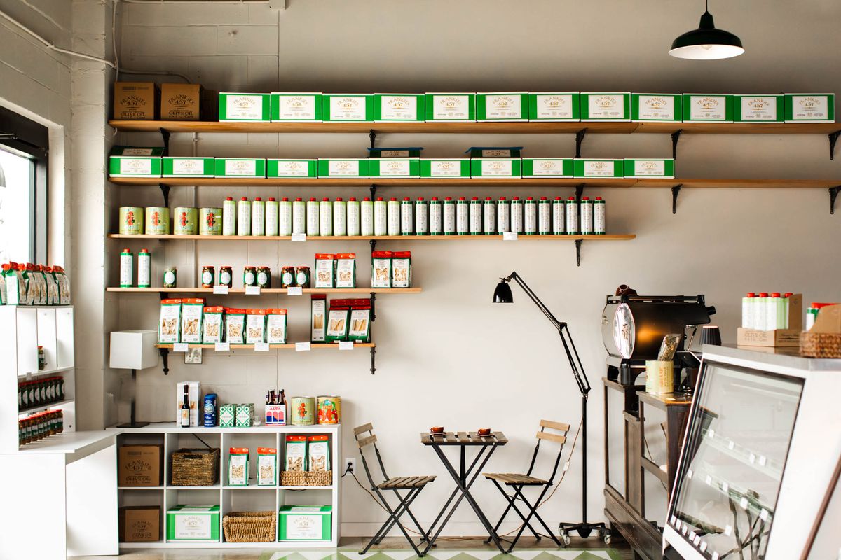 A minimalist market with several shelves stocked with green boxes and cans of olive oils and preserves. There’s a table for two in the enter and a salumi case to the right of the frame.