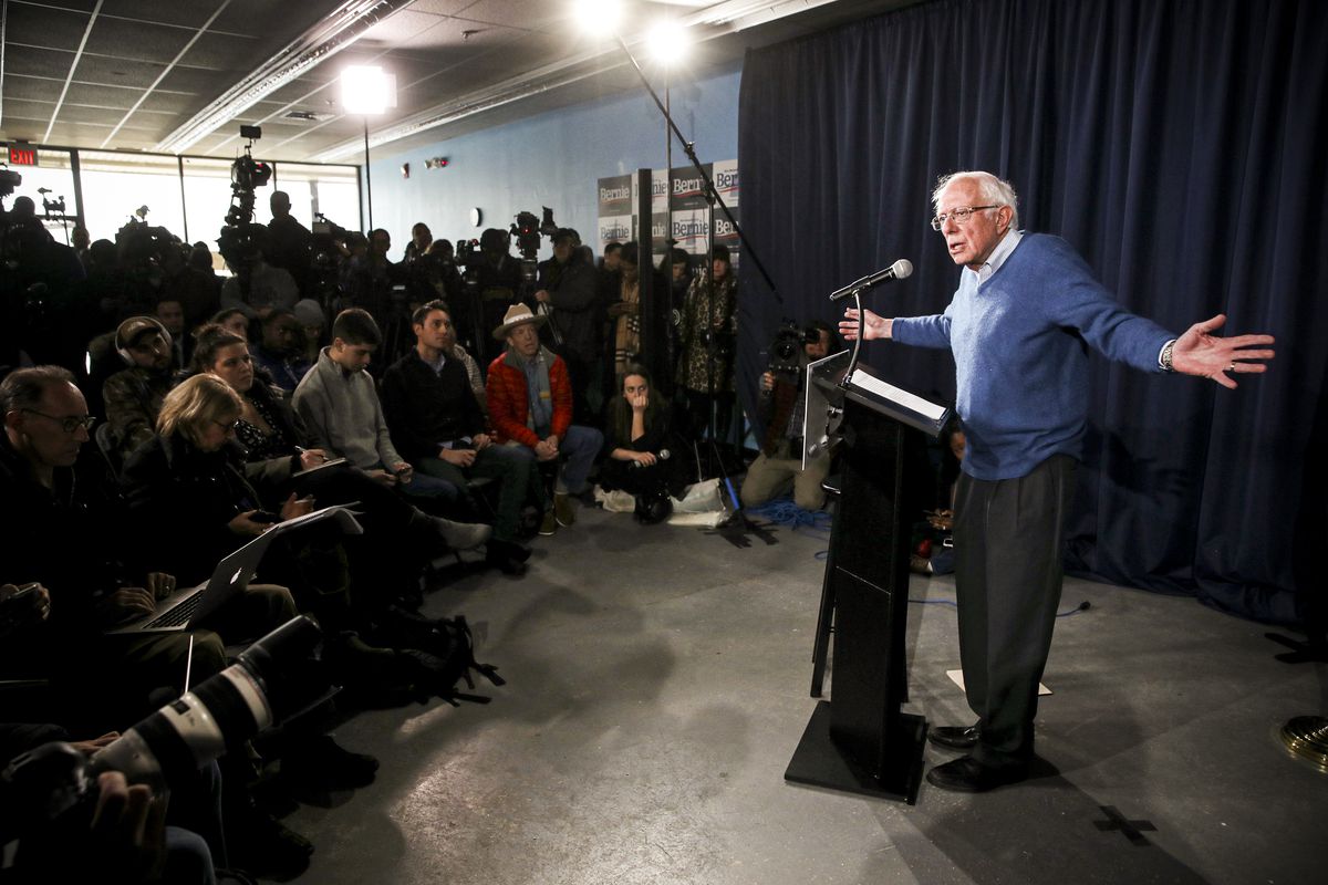 Senator Bernie Sanders standing at a podium, speaking into a microphone, with his arms out wide.