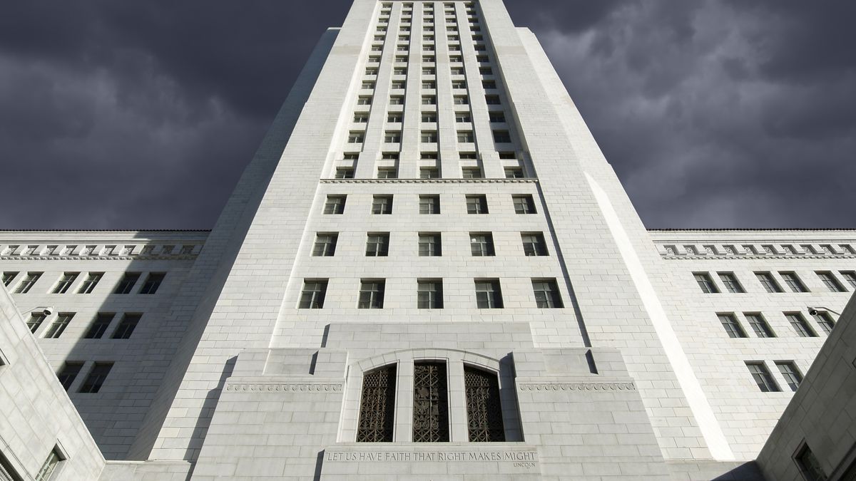 A worms-eye view of a looming white concrete building, L.A. City Hall, many stories high. It feels ominous and foreboding, towering above the viewer’s vantage point.
