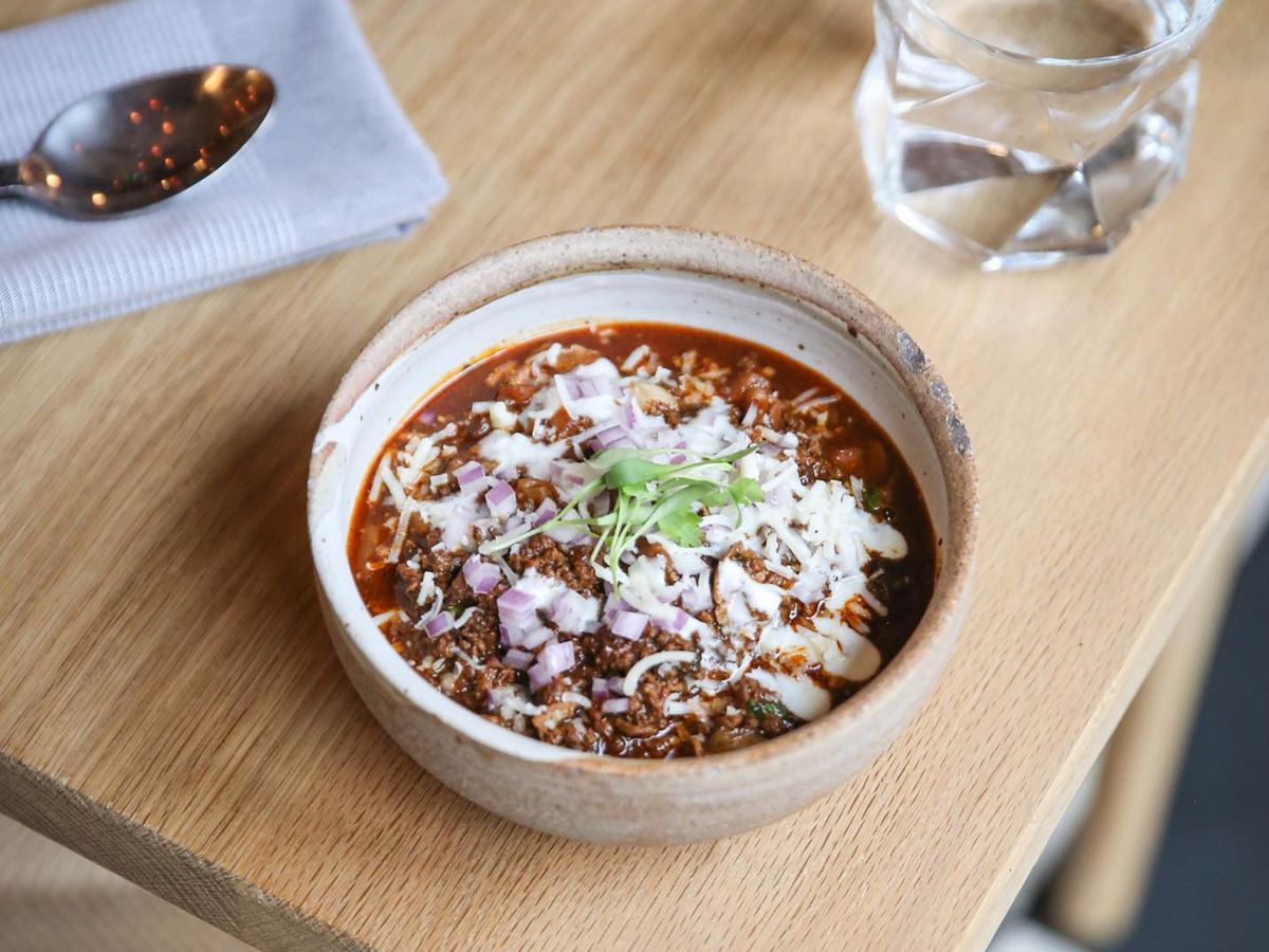 A bowl of chili topped with white cheese on a wooden table with a silver spoon to the left.