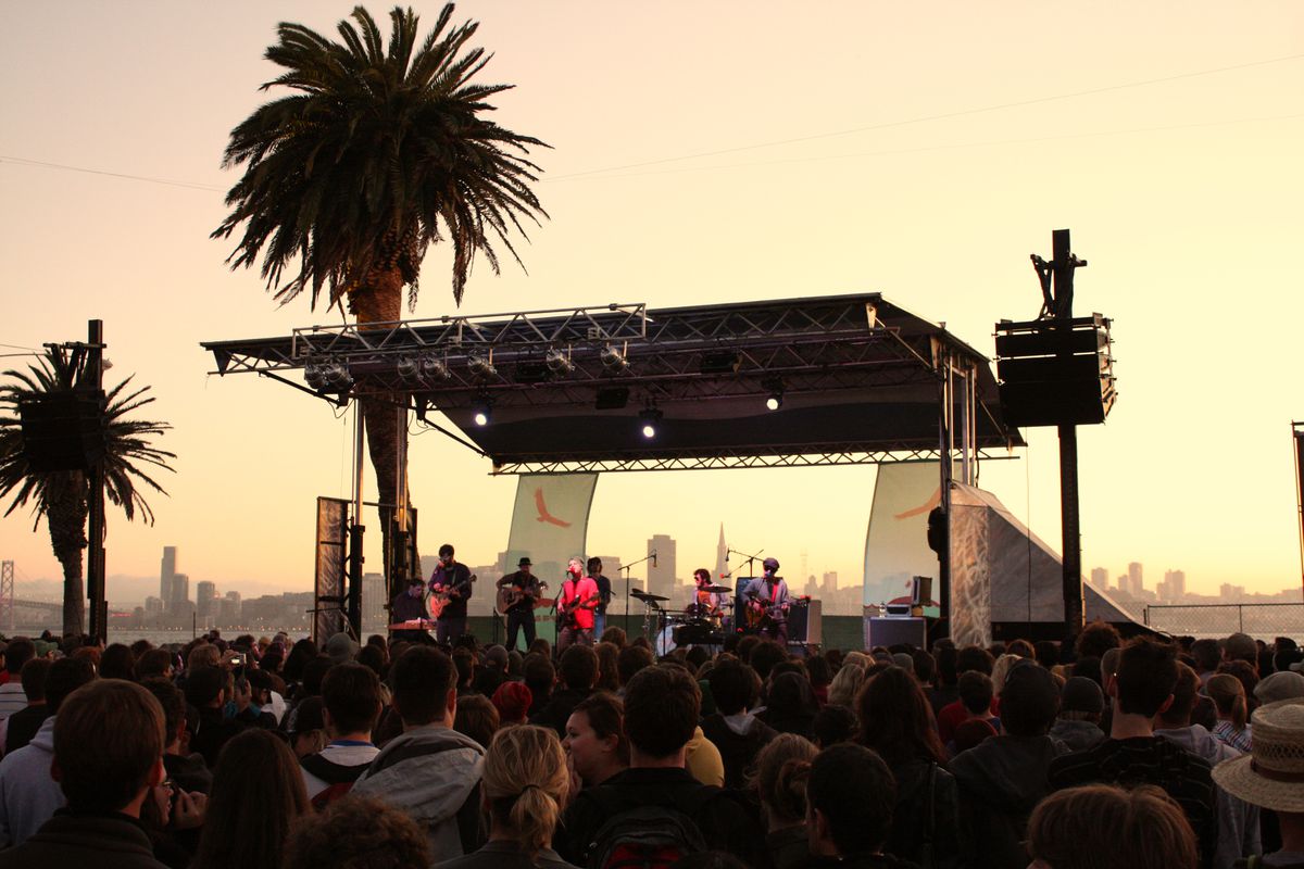 A band playing onstage at Treasure Island, with the San Francisco skyline silhouetted in the sunset behind them.