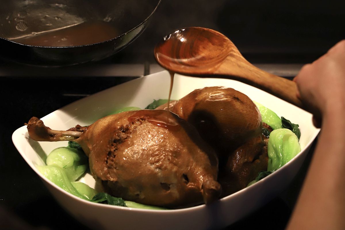 A deboned, stuffed, and crisped eight treasure duck sits on a bed of pak choi, as a cook spoons over its cooking juices