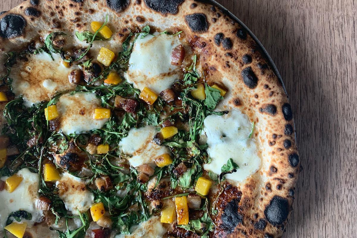 A pizza with a charred crust topped with melted white cheese, yellow corn kernels, and green leaves.